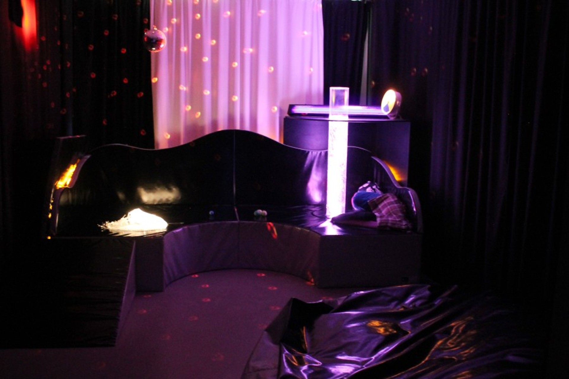Dunkler Snoezelen-Raum mit lila Leuchteffekten: Eine sprudelnde Lichtsäule steht neben einem geschwungenen schwarzen Polsterpodest. Spiegelkugel und Lichtpunkte beleuchten den Raum.