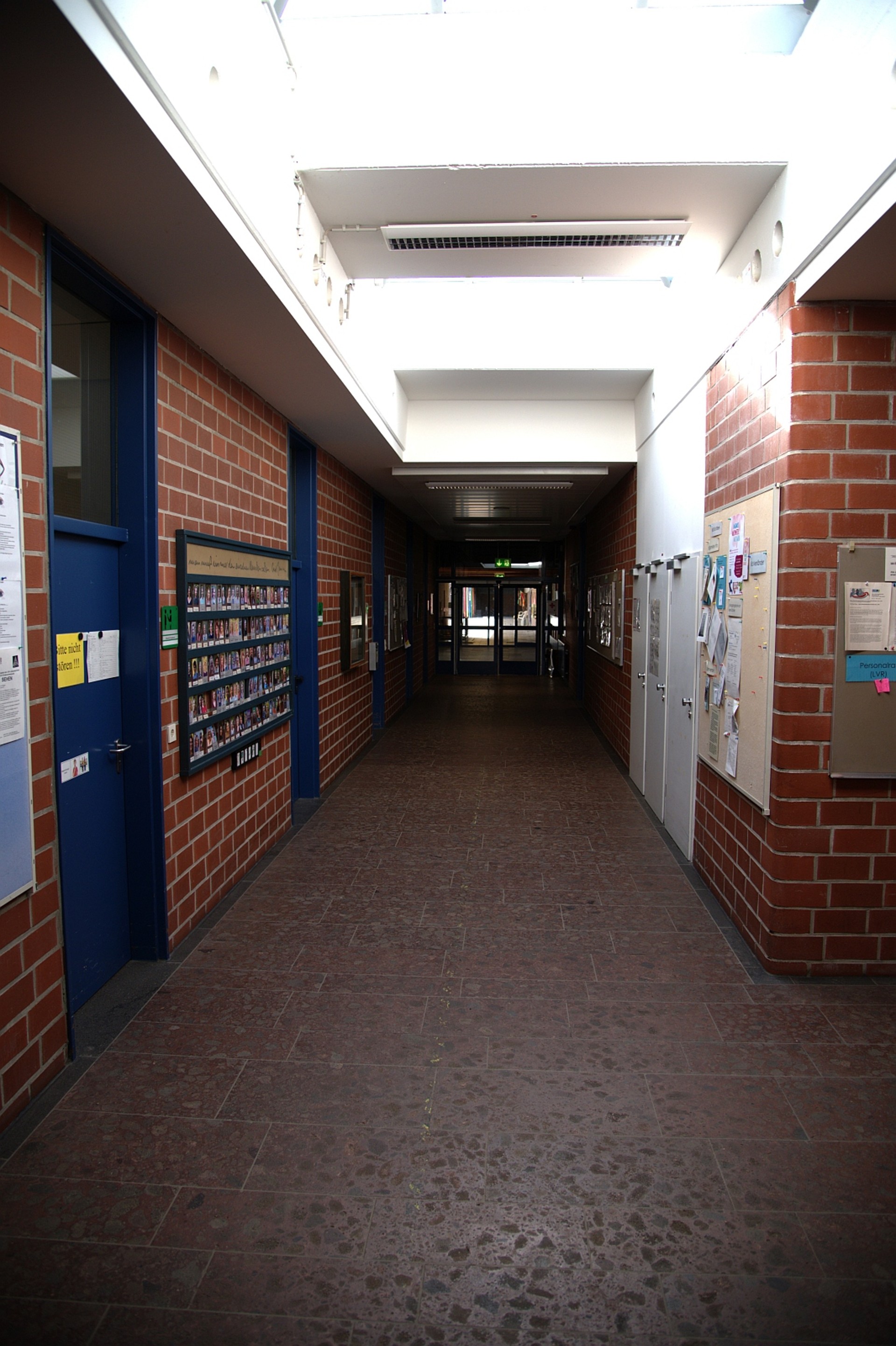 Ein langer, heller Schulflur mit rötlichem Steinboden und Sichtmauerwerk aus Backstein. Große Oberlichter lassen viel Tageslicht herein. An den Wänden hängen Infotafeln und Pinnwände; blaue Türen führen in die angrenzenden Klassenräume.