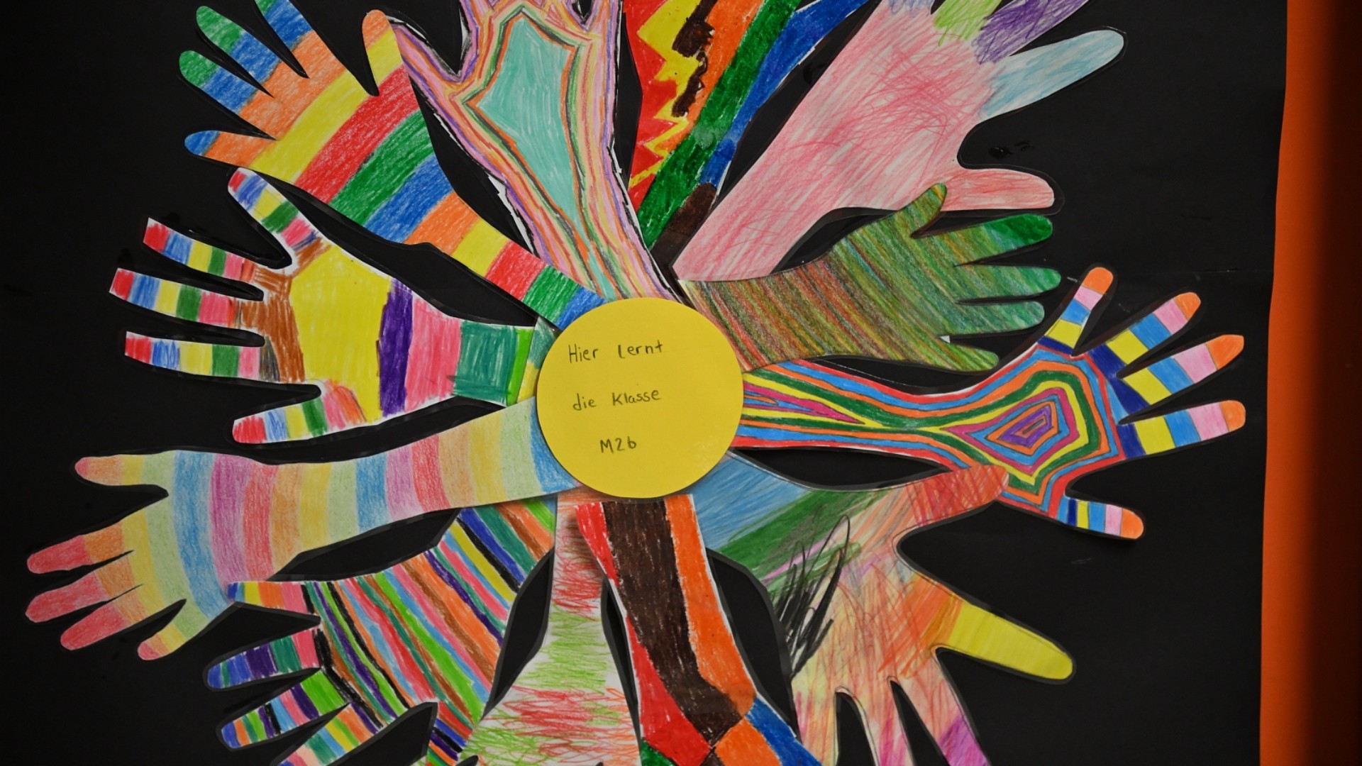 Im Vordergrund einer roter Sandplatz für Fußball und Basketball, im Hintergrund ein Hof mit Bänken, einem grün-bunt bemalten Holzhäuschen und ganz hinten das zweistöckige Schulgebäude mit grünen Fensterrahmen.