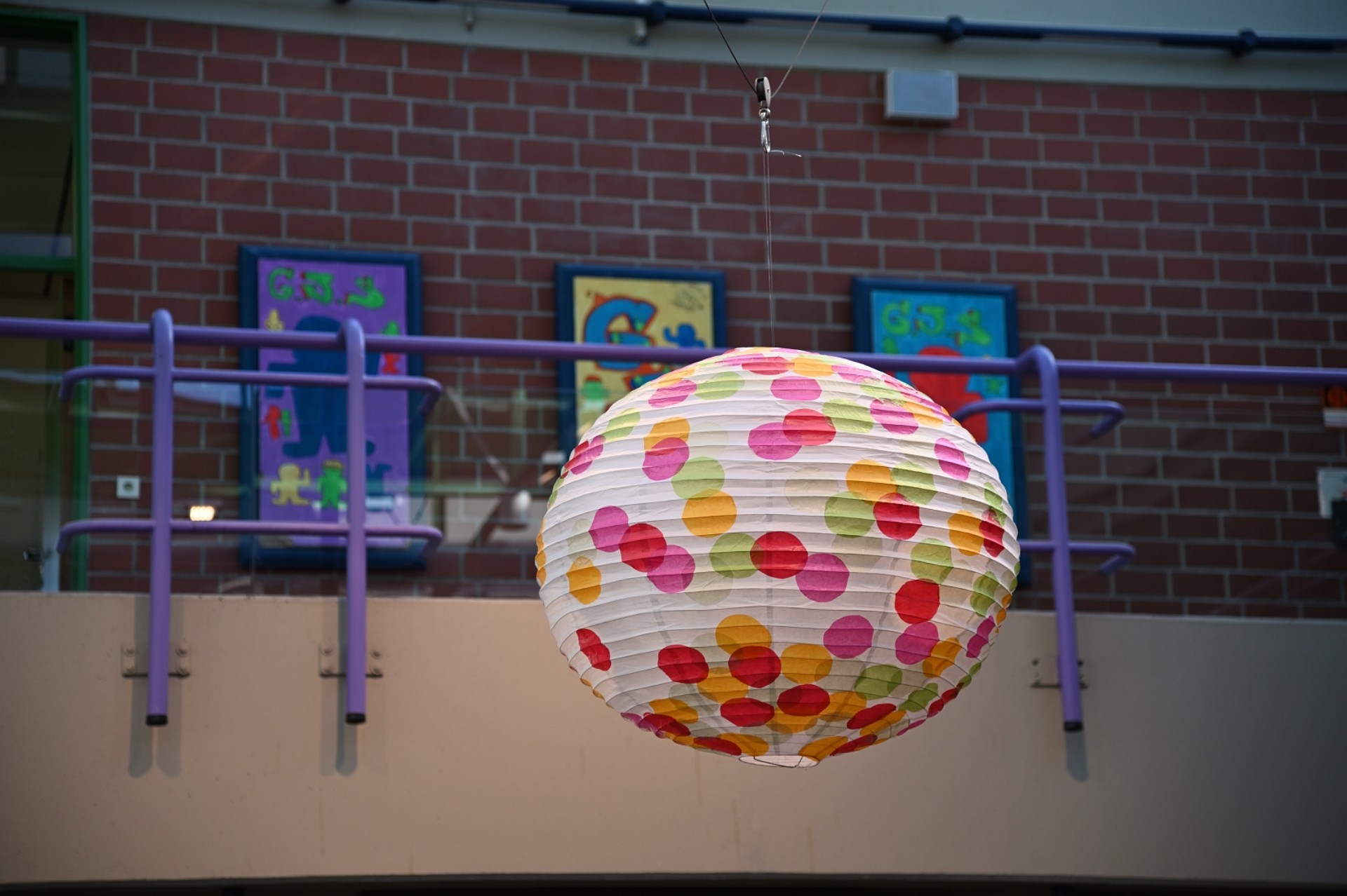 Eine große, runde Papierlaterne mit bunten Punkten hängt an einem dünnen Draht im offenen Bereich eines Treppenhauses. Im Hintergrund sind ein lilafarbenes Geländer und bunte Schülerarbeiten an einer Backsteinwand zu sehen.