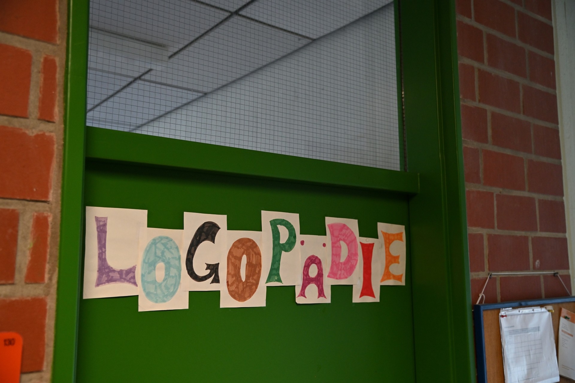 Detailansicht einer grünen Tür, auf der das Wort „LOGOPÄDIE“ in bunten, individuell gestalteten Papierbuchstaben aufgeklebt ist.