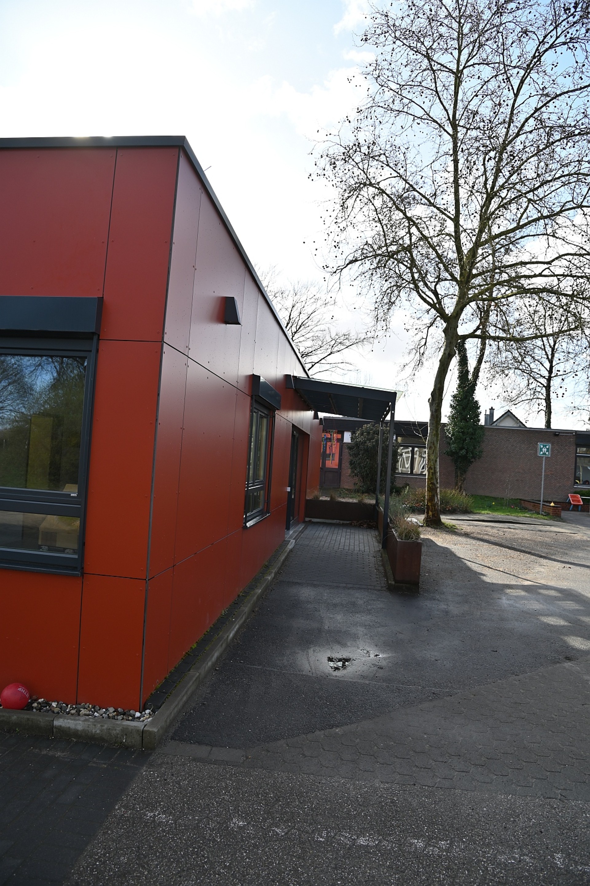 Die Außenansicht eines modernen, flachen Gebäudes mit einer markanten roten Fassadenverkleidung. Vor dem Haus befindet sich ein asphaltierter Weg, und im Hintergrund ragen hohe, noch weitgehend kahle Bäume in den leicht bewölkten Himmel.