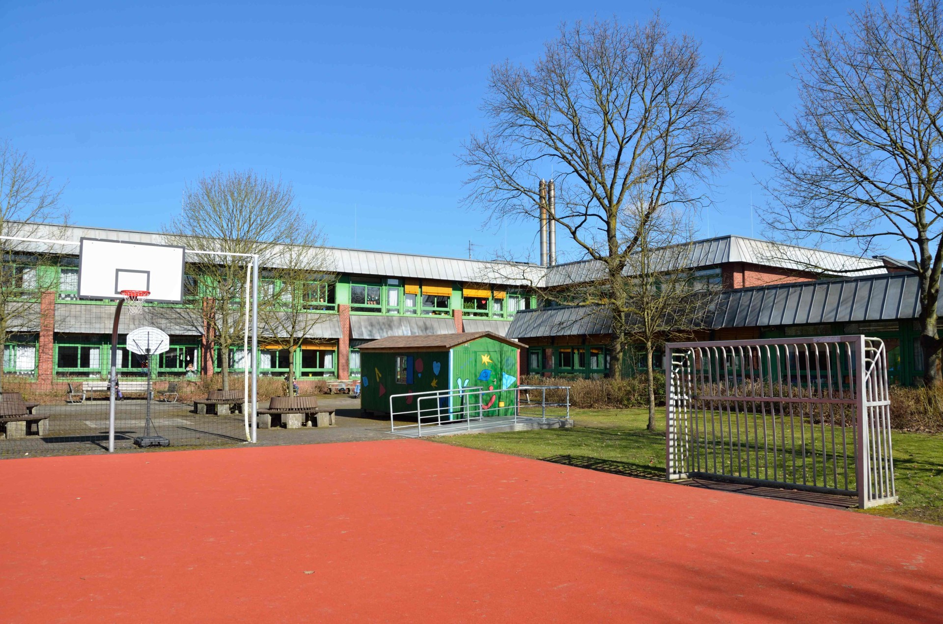 Im Vordergrund einer roter Sandplatz für Fußball und Basketball, im Hintergrund ein Hof mit Bänken, einem grün-bunt bemalten Holzhäuschen und ganz hinten das zweistöckige Schulgebäude mit grünen Fensterrahmen.