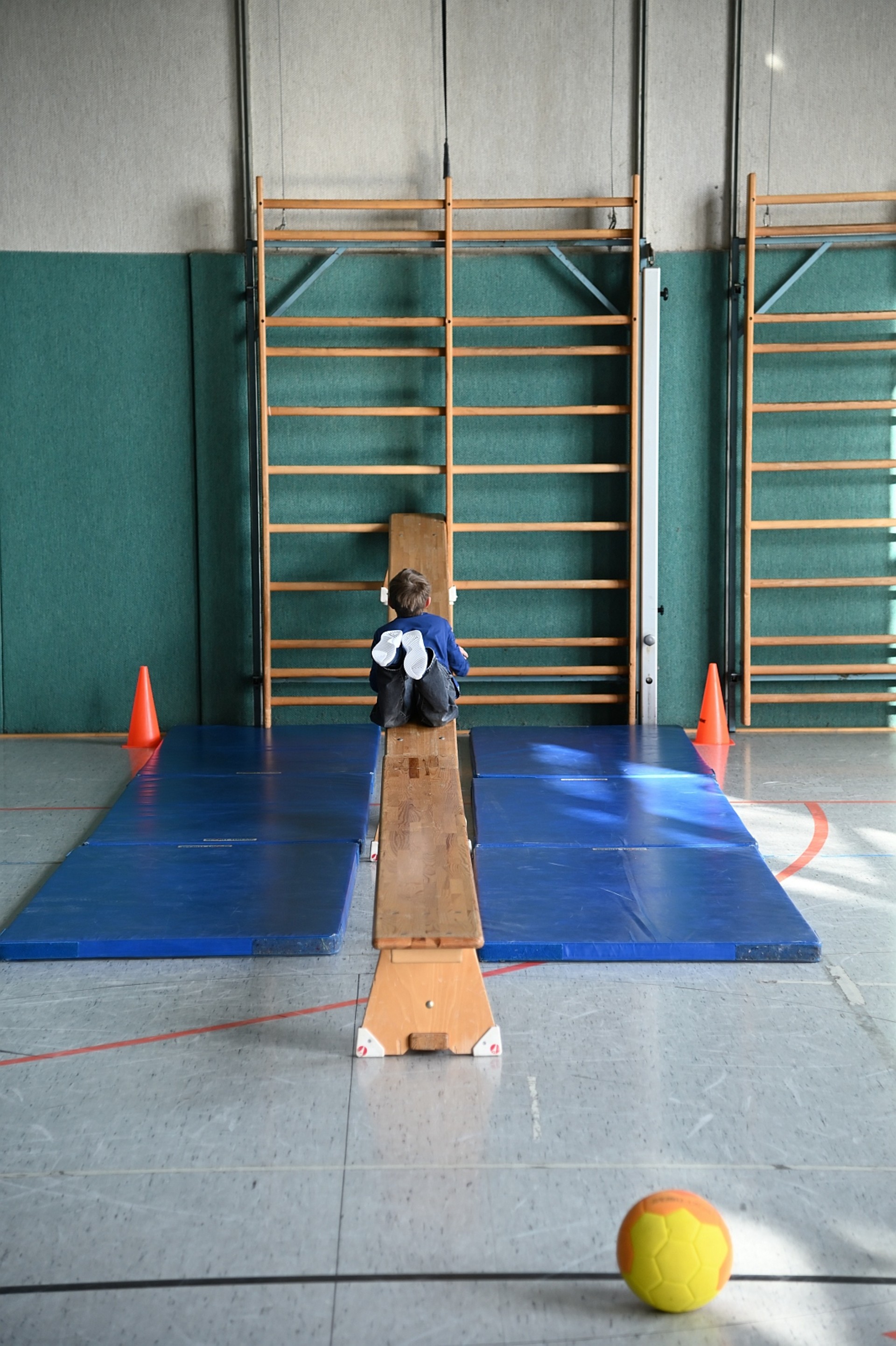 Blick in eine Sporthalle. Ein Kind liegt bäuchlings mit dem Rücken zur Kamera auf einer schräg an einer Sprossenwand eingehängten Turnbank. Blaue Matten sichern den Bereich ab, und im Vordergrund liegt ein gelb-orangefarbener Ball auf dem Hallenboden.
