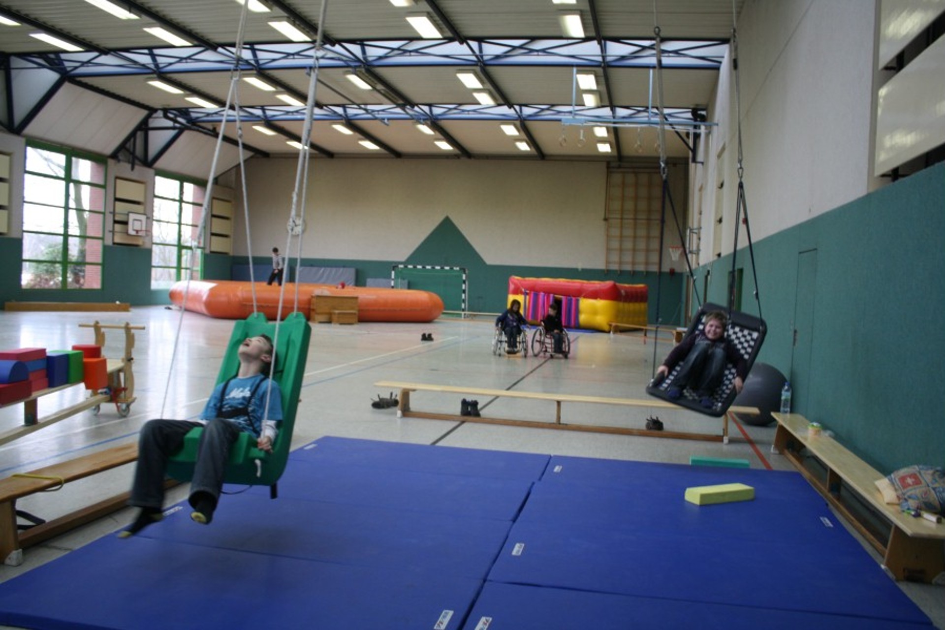 Große Turnhalle mit Inklusions-Sportgeräten: Kinder nutzen spezielle Hängeschaukeln über blauen Matten. Im Hintergrund sind Kinder im Rollstuhl sowie eine Hüpfburg und eine große Luftmatte zu sehen.