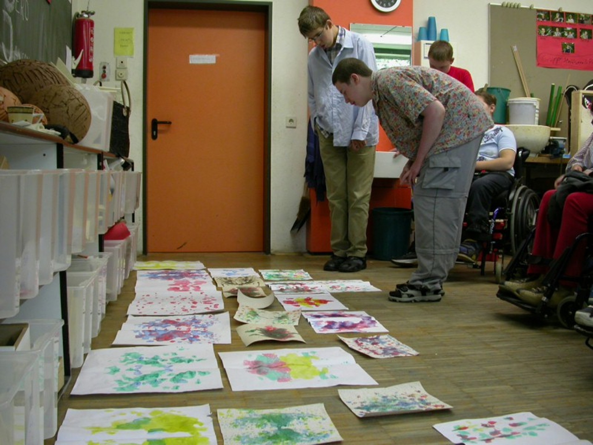 In einem Schulraum liegen mehrere bunte Bilder auf dem Boden, zwei Jugendliche beugen sich darüber. Im Hintergrund sind weitere Kinder zu erahnen, die teilweise im Rollstuhl sitzen.