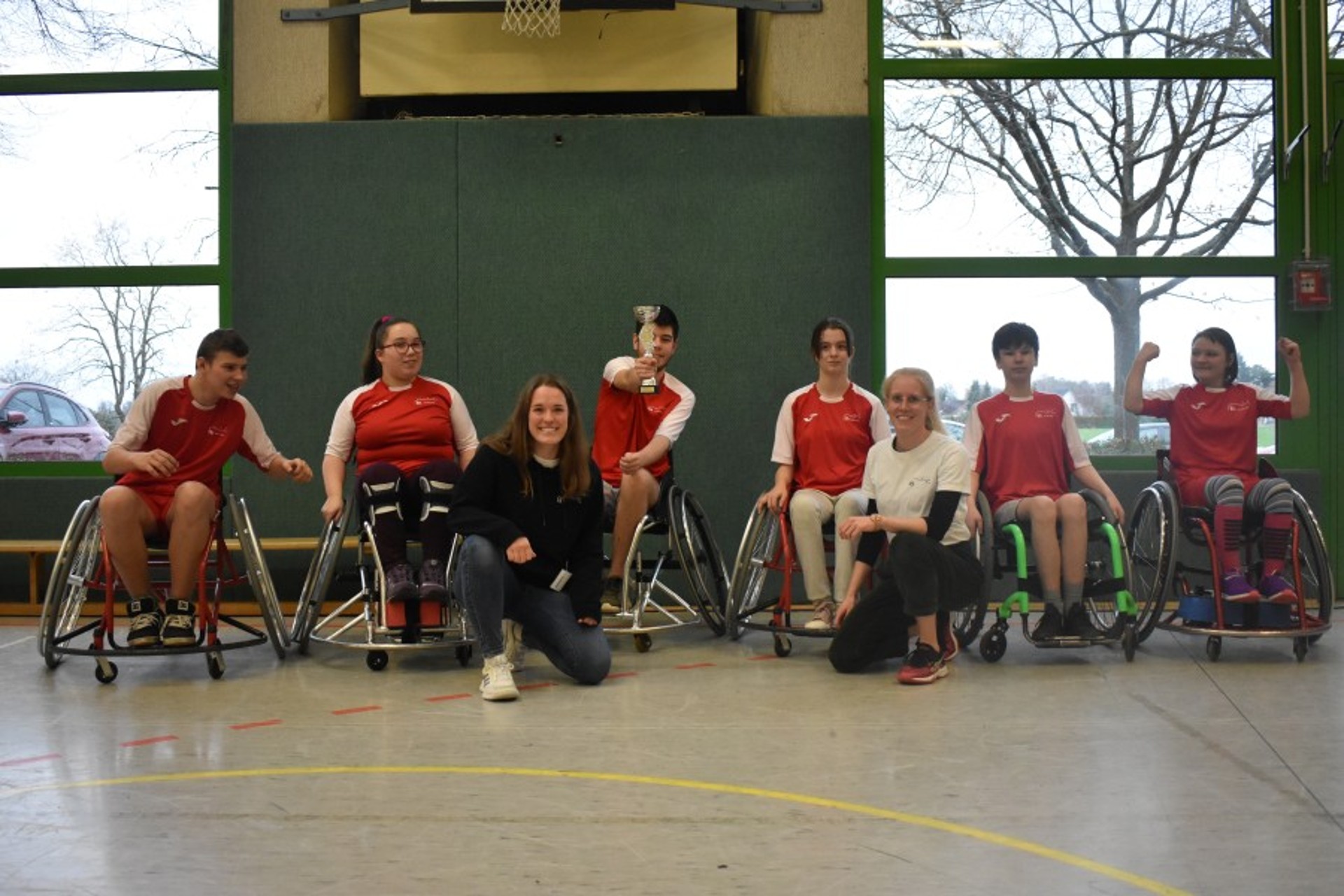 Ein Team aus sechs Rollstuhl-Basketball-Spieler*innen mit roten Krefels-Trikots berät sich mit seinen Trainerinnen im Kreis.