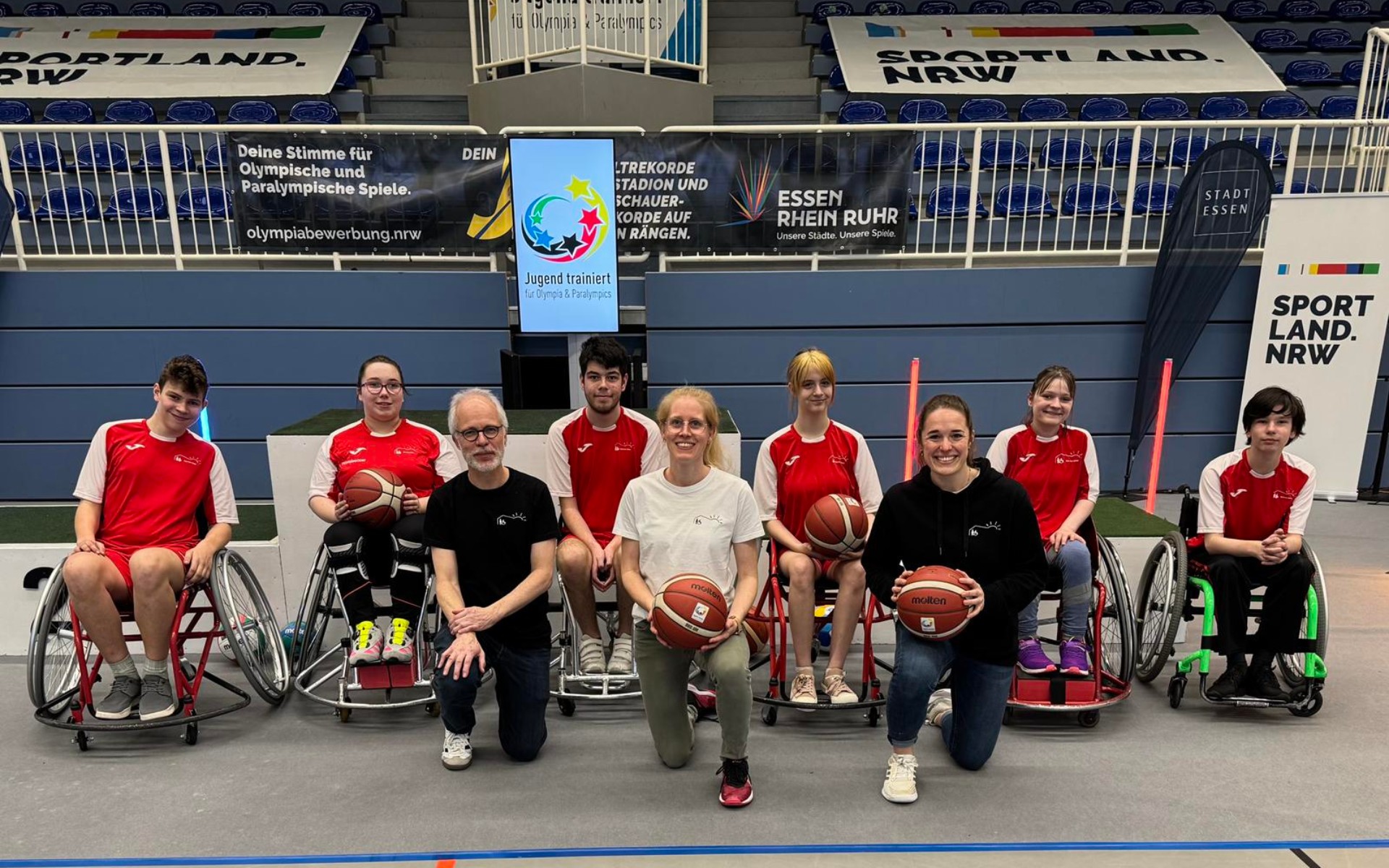 Ein Team aus Rollstuhl-Basketball-Spieler*innen mit roten Krefels-Trikots und deren TrainerTrainerinnen im Kreis.