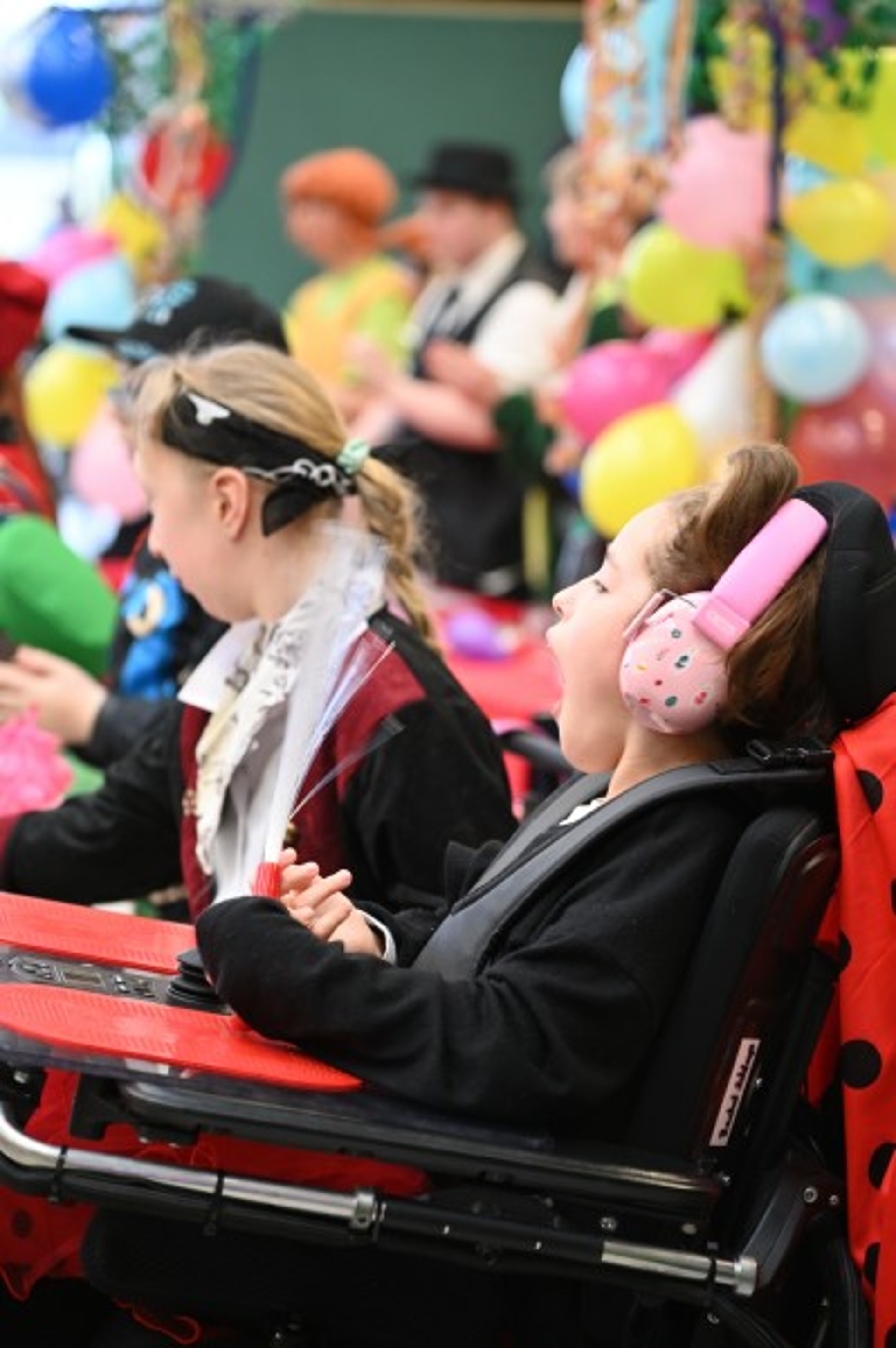 Zwei verkleidete Kinder im Rollstuhl schauen auf buntes Karnevalsgeschehen