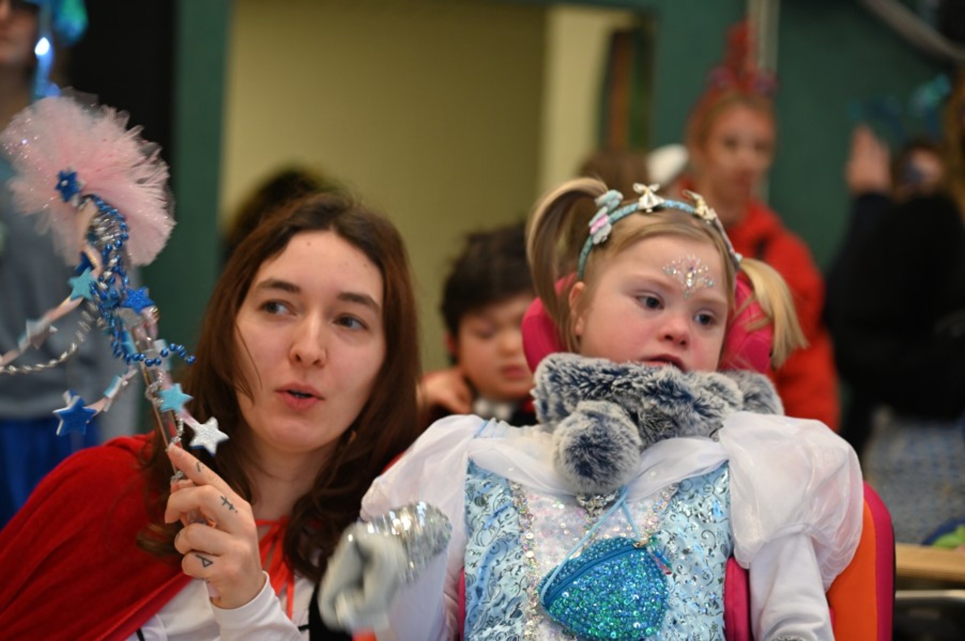 Ein Mädchen im Rollstuhl ist als Eiskönigin verkleidet. Neben ihr schwenkt eine Frau mit roten Umhang einen Feenstab mit vielen blauen Sternen.