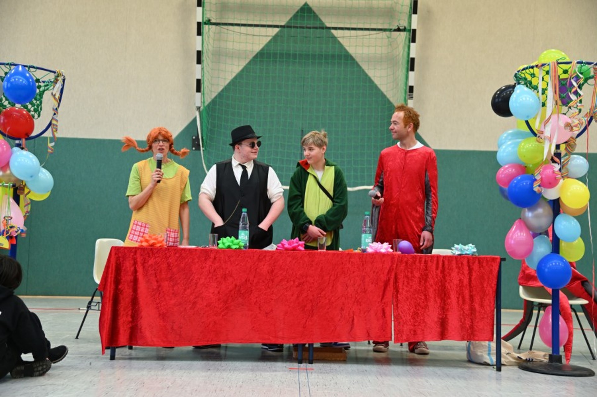 Blick in eine bunt geschmückte Turnhalle: vorne steht ein Tisch mit einer roten Samtdecke. Hinter ihm stehen Pipi Langstrumpf, einer von den Blues Brothers, ein Dinosaurier und das Sams.