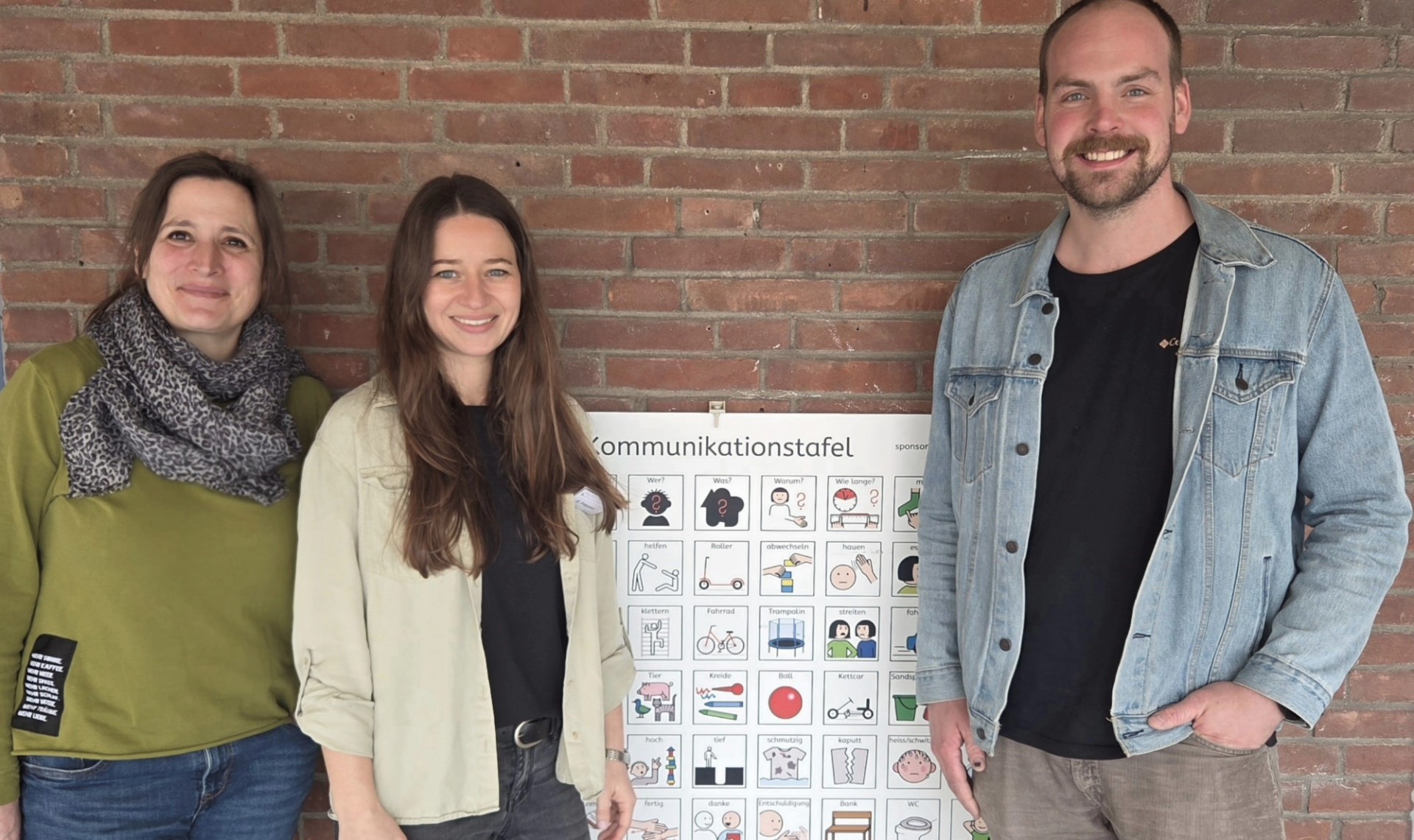 Die zwei Lehrerinnen und der Lehrer des Teams Unterstützte Kommunikation stehen vor einer gemauerten Wand mit einer Kommunikationstafel mit Metacom-Symbolbildern