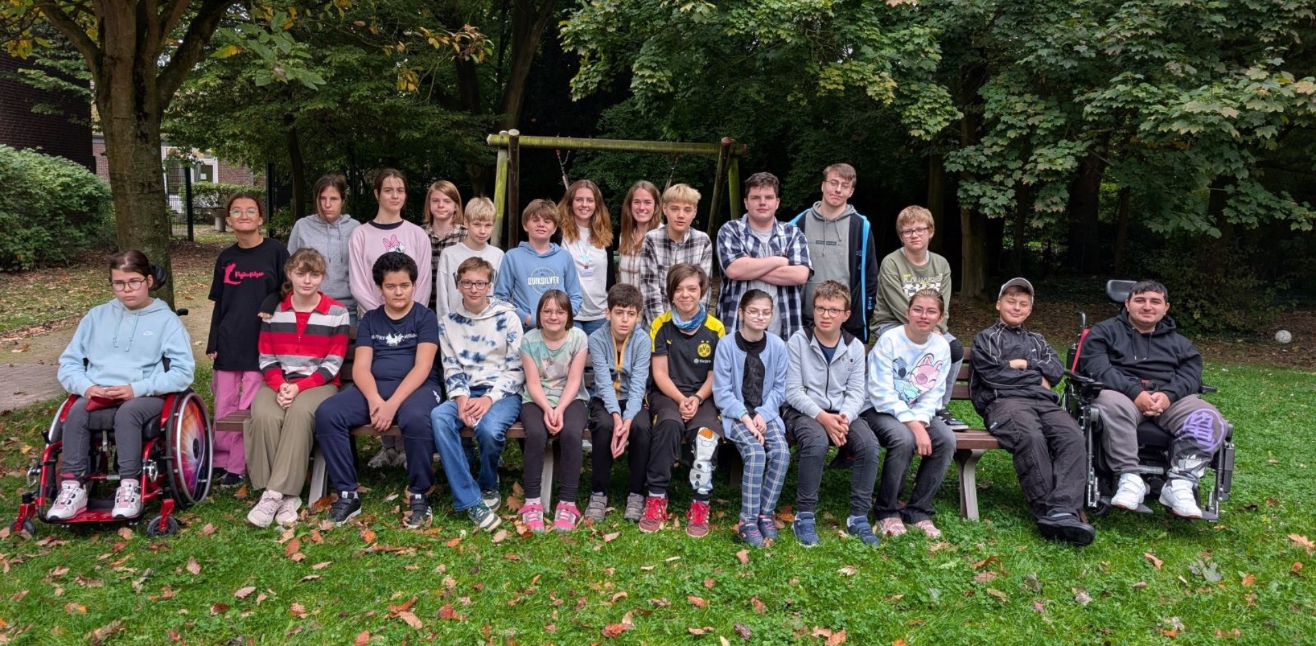 Etwa 20 Teenager haben sich in zwei Reihen um eine Bank im Grünen zum Gruppenfoto postiert. Zwei Jugendliche sitzen im Rollstuhl. In der Mitte der hinteren Reihe stehen auch zwei Lehrerinnen. Alle lächeln freundlich.