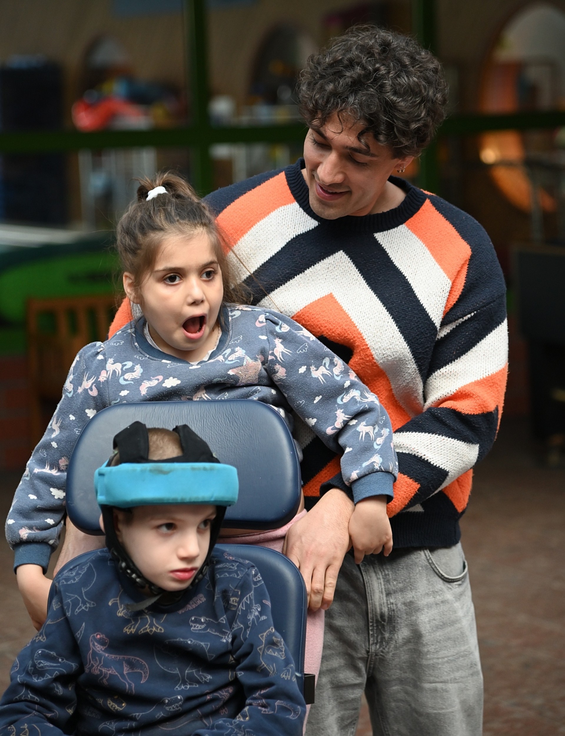 Ein junger Mann hält ein Mädchen vor sich, das bei einem weiteren Kind hinten auf dem Rollstuhl steht. Dieses Kind trägt einen blauen Kopfschutz.