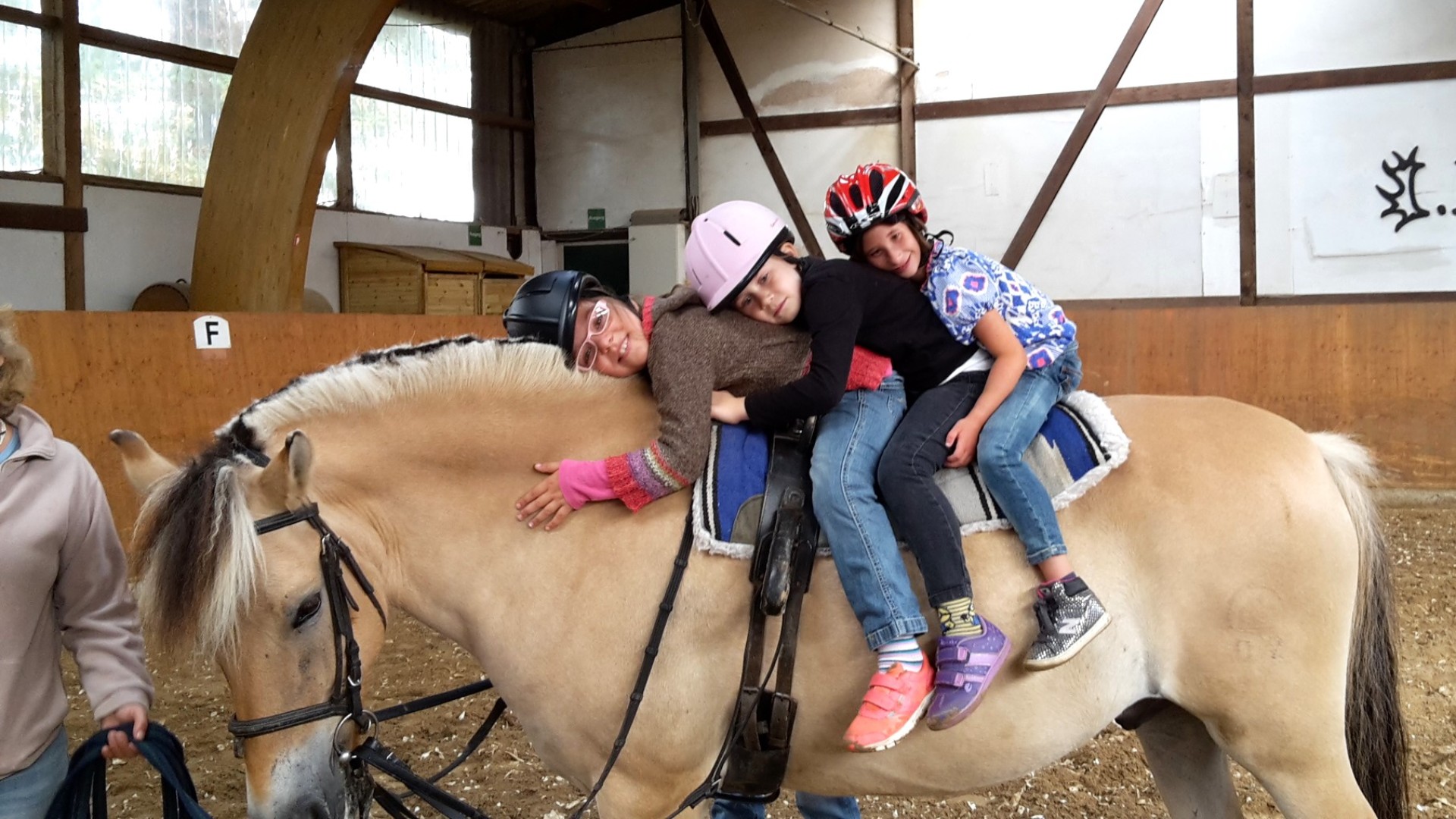In einer Reithalle sitzen drei Mädchen mit Helmen hintereinander auf einem Pferd. Sie lehnen sich nach vorne, so dass jeweils ein auf der anderen lehnt. Das Pferd wird an einer Leine geführt.